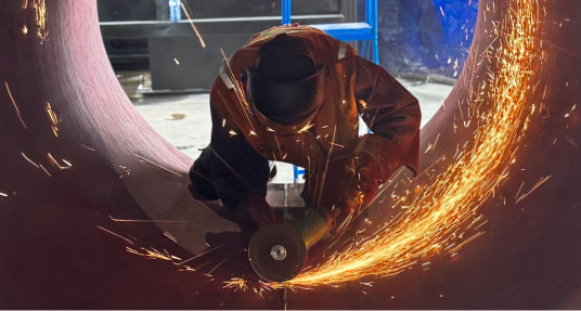 A welder in full protective gear crouches beside a large metal pipe, grinding its surface with an angle grinder